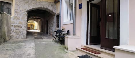 A narrow alleyway with a stone archway leading to a building with a dark wooden door and a small outdoor seating area with a table and chairs.