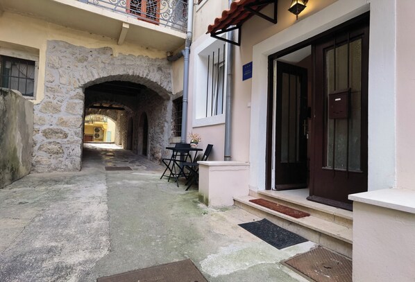 Une ruelle étroite, surmontée d’une arche en pierre, mène à un bâtiment doté d’une porte en bois sombre et d’un petit espace extérieur aménagé avec une table et des chaises.