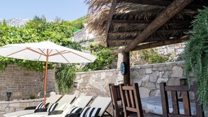 A sunlit outdoor lounge area with lounge chairs and a table, surrounded by stone walls and greenery. - House Wood (Orebic)