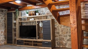 A rustic wooden entertainment unit with a flat-screen TV, shelves, and drawers against a stone wall. - House Wood (Orebic)