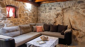 A cozy room with a stone wall, a cushioned sofa, a small table with a bowl, and a window with checkered curtains. - House Wood (Orebic)