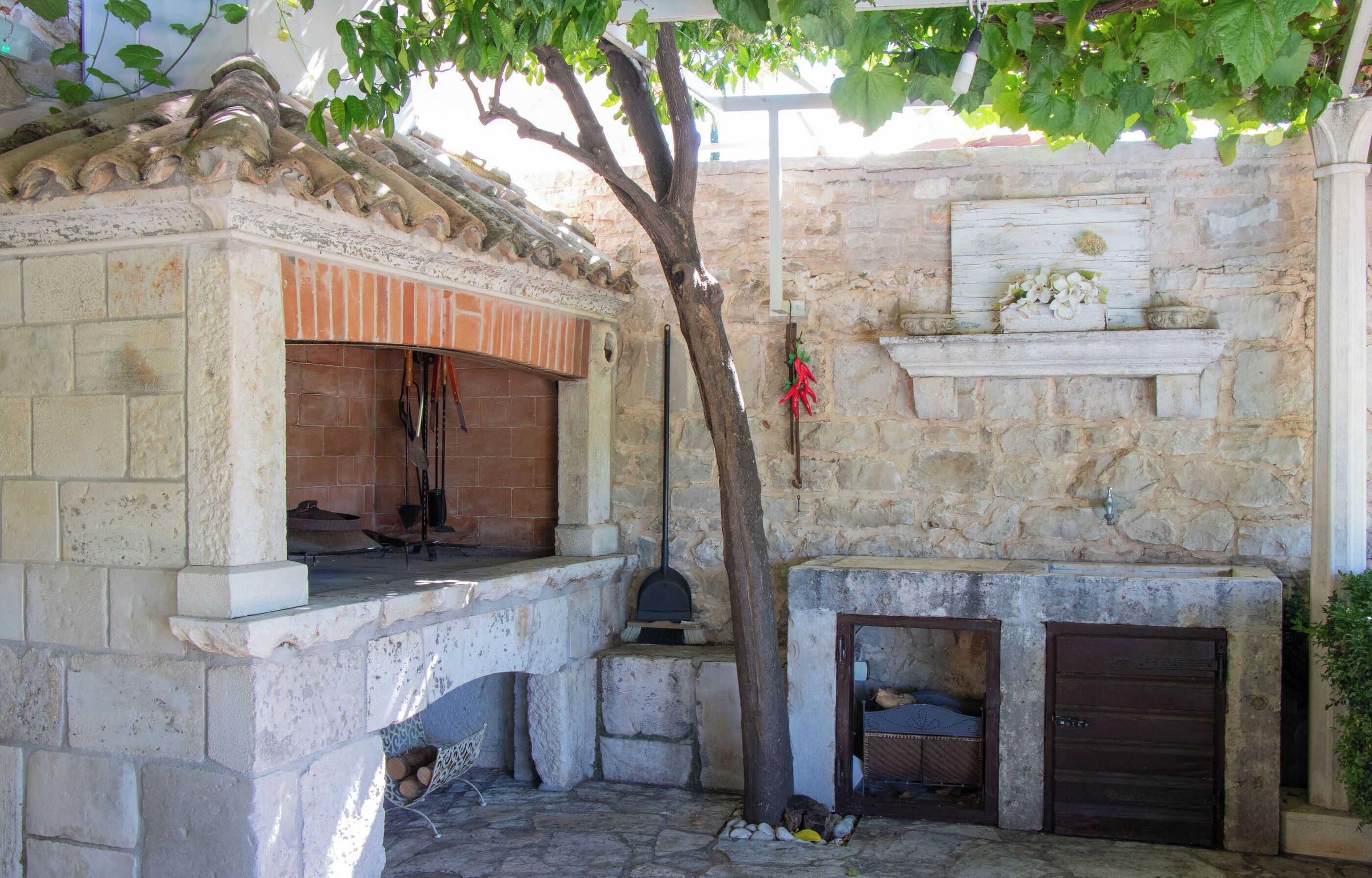 Cocina exterior de piedra con chimenea, un árbol y una pared de piedra con repisa.