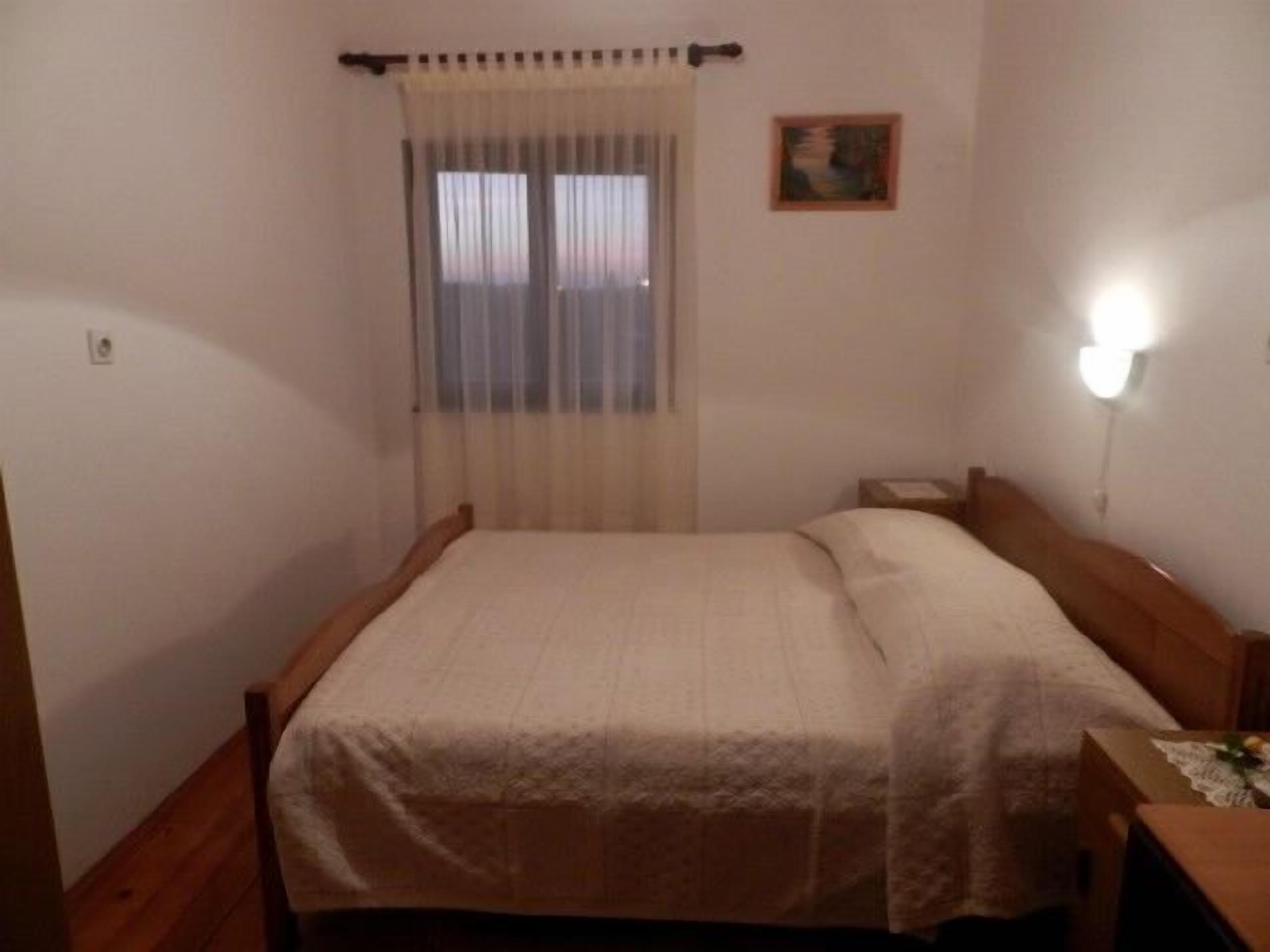 A single bed with a wooden headboard, a bedside table, a nightstand, a window with sheer curtains, and a wall-mounted light.