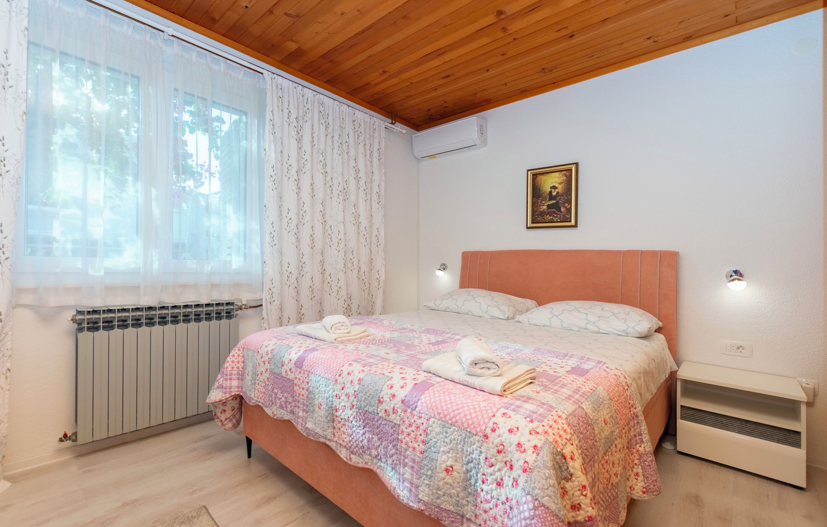 Een slaapkamer met een houten plafond, een bed met een quilt met bloemenprint, een radiator en een wandmodel airconditioner.