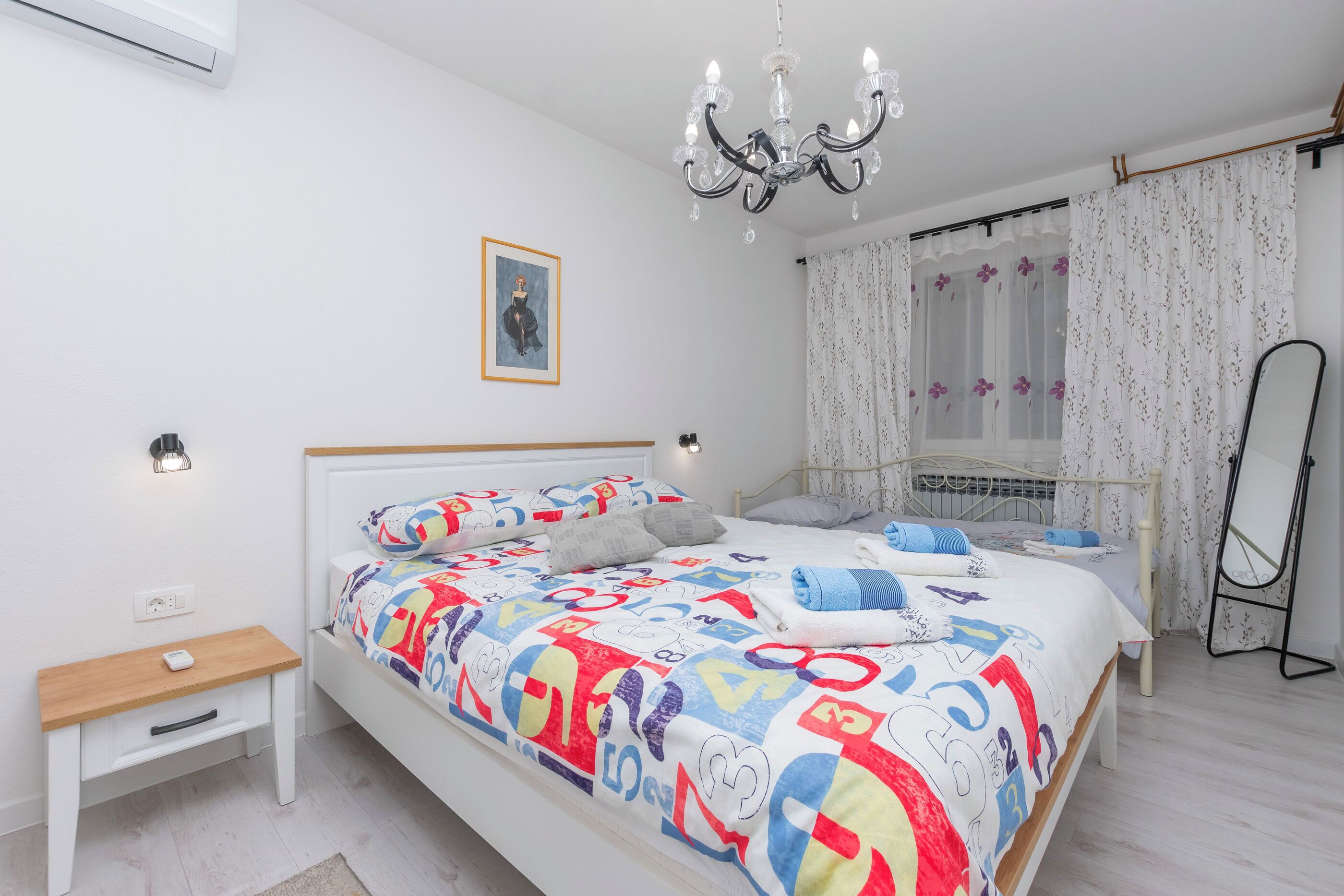 A bedroom with a bed, bedside table, nightstand, and a standing mirror.