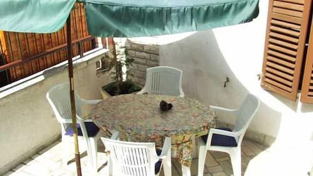 Ein runder Tisch mit einer blumigen Tischdecke, umgeben von weißen Stühlen, unter einem grünen Sonnenschirm.