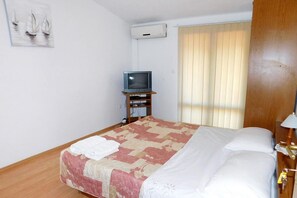 A hotel room with a bed, a television on a wooden stand, a window with blinds, and a wall-mounted air conditioner. - Apartments Ljiljana (Smokvica)