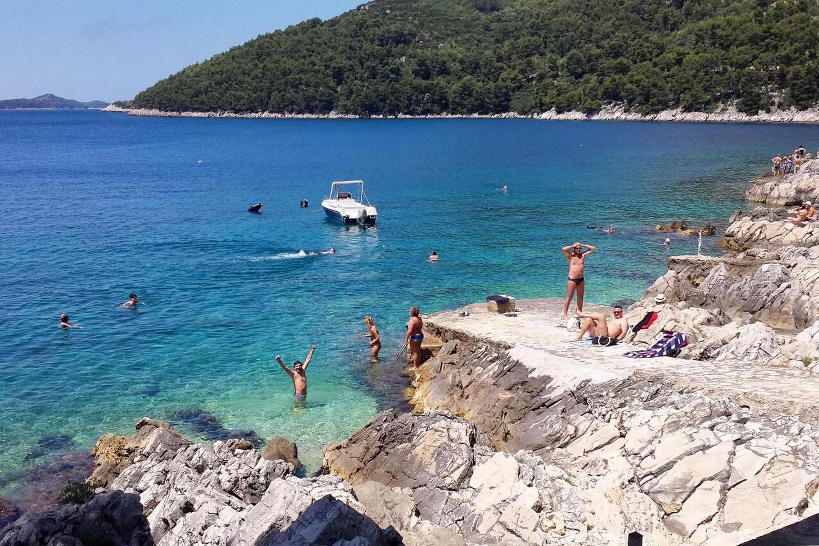 Uma praia rochosa com água azul cristalina, pessoas nadando e tomando sol, e um barco ao longe.