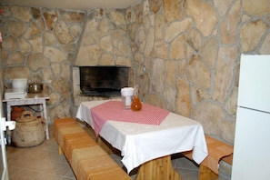 A stone-walled room with a fireplace, a table with a checkered tablecloth, benches, and a small table with kitchen items. - Apartments Ljiljana (Smokvica)