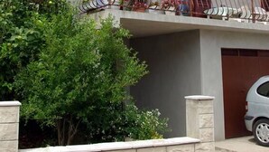 Ένα διώροφο κτίριο με μπαλκόνια, ένα αυτοκίνητο σταθμευμένο στην αυλή και ένας πέτρινος τοίχος στο προσκήνιο.