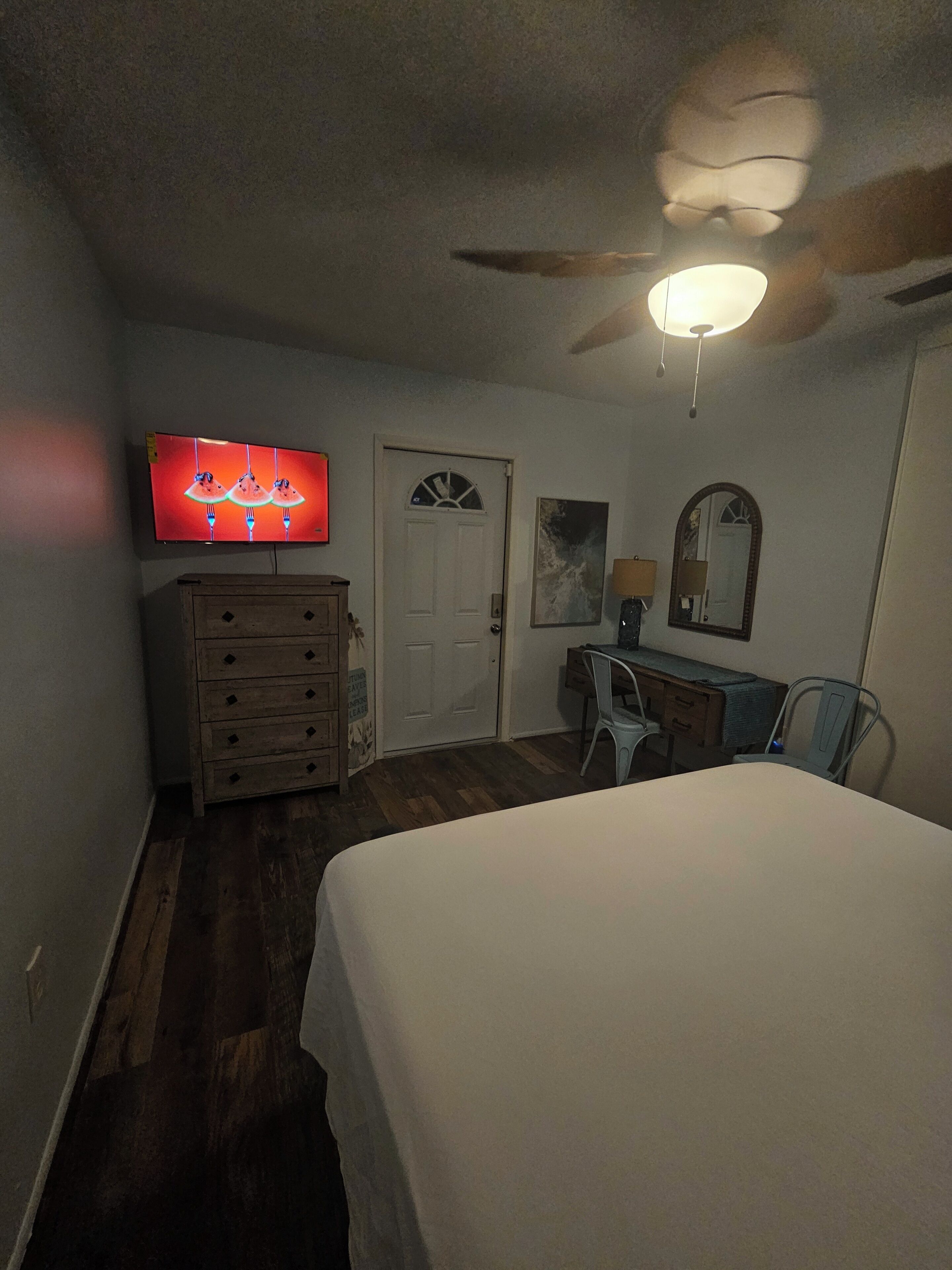 Une chambre à coucher comprenant un lit, une commode, un bureau avec une chaise, un ventilateur de plafond et un téléviseur fixé au mur.
