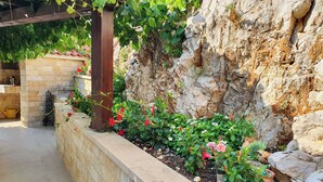 Jardim com muro de pedra, diversas plantas e flores, pérgola coberta por trepadeiras e um muro de pedra rústico.