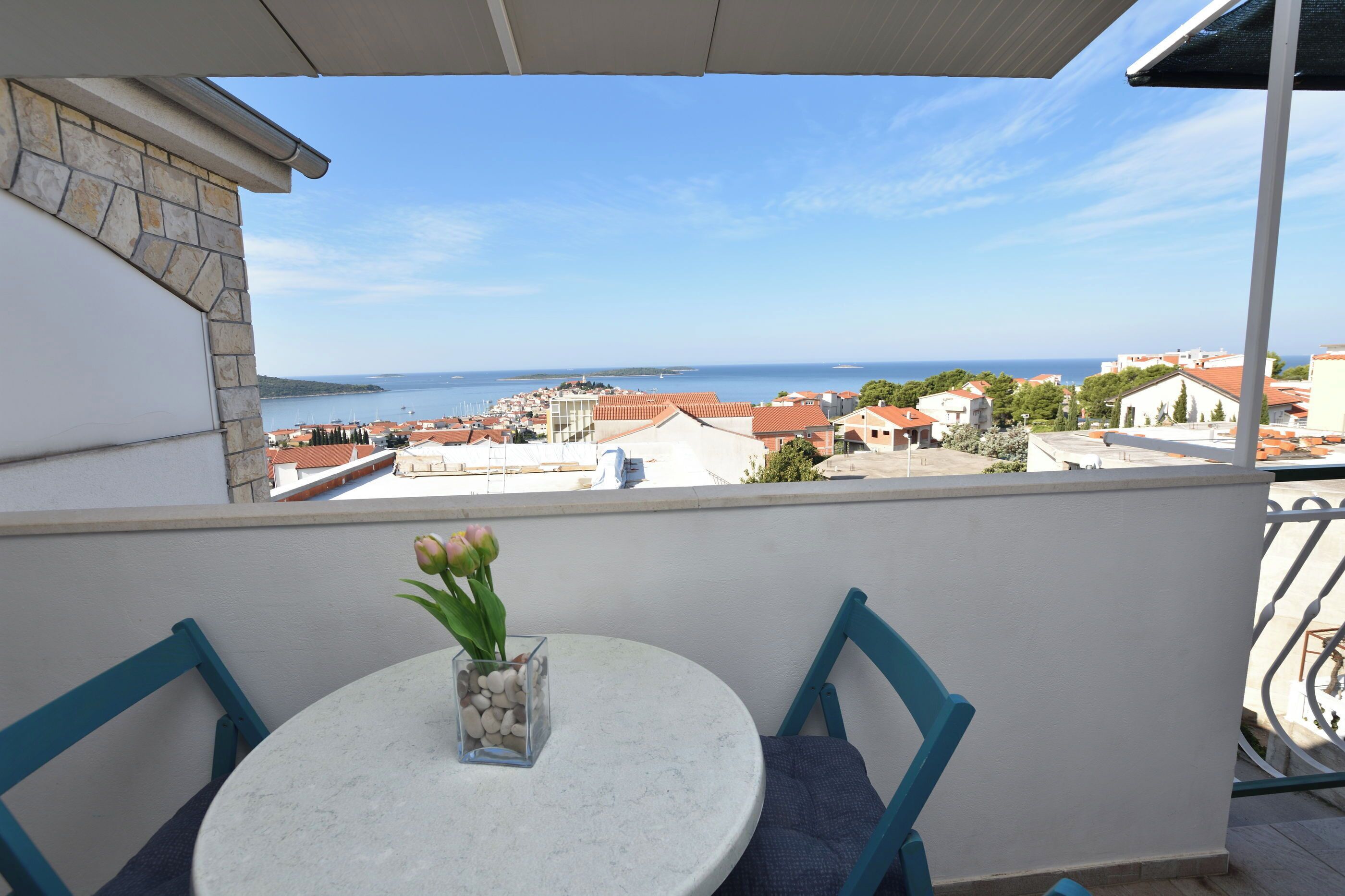 A balcony with a table and chairs, overlooking a coastal town with buildings and the sea.