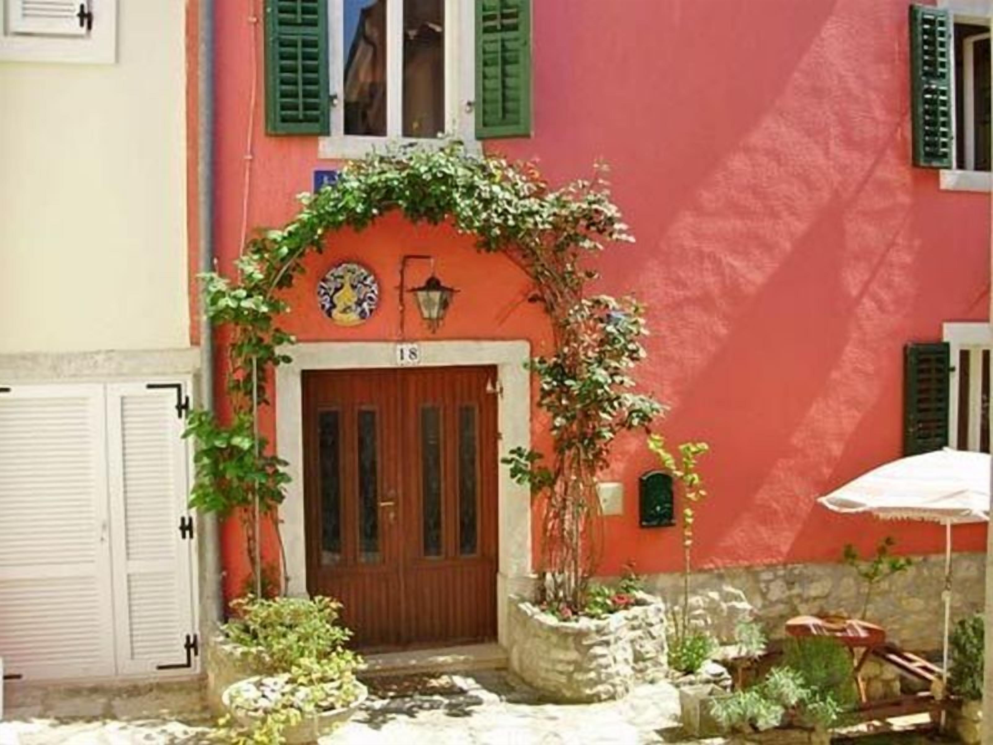 Un edificio rojo con puerta de madera, contraventanas verdes y un cartel con el número 18 sobre la puerta.