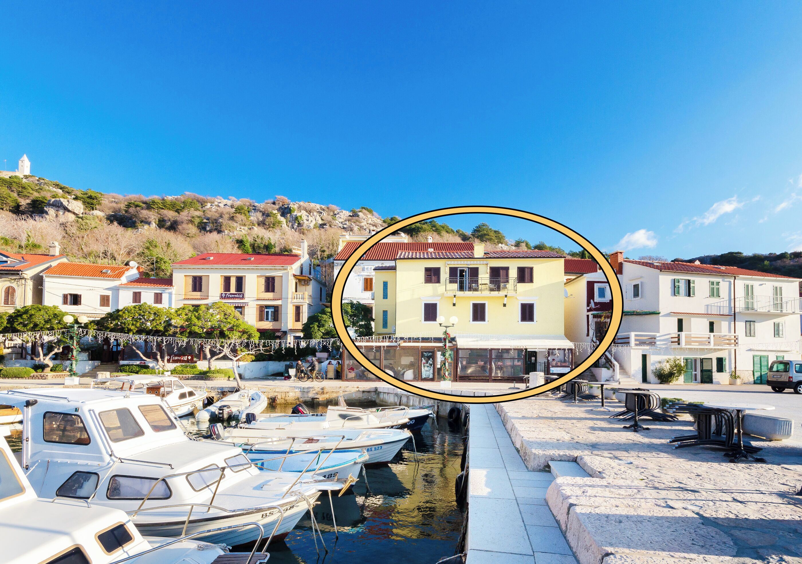 A harbor with boats docked, a building with a red roof, and a hill in the background.