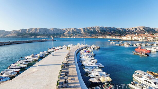 A marina with boats docked along a curved pier, a clear sky, and a mountainous backdrop. - Apartment Jasna By the sea (Baska)