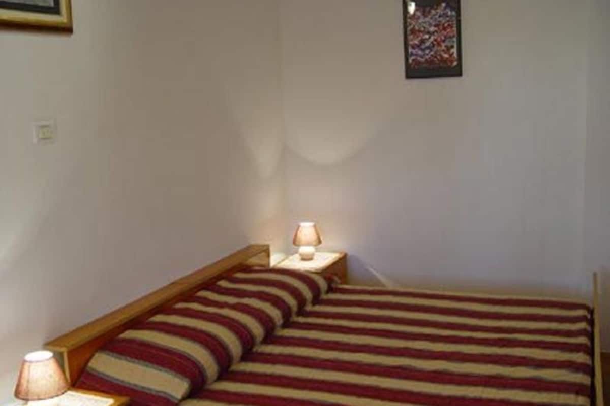 A small, single-bed room with a striped bedspread, two bedside lamps, and a framed picture on the wall.