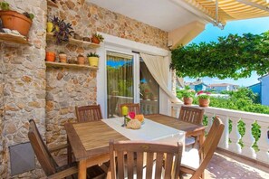 Un balcon aux murs de pierre, agrémenté d’une table et de chaises en bois, de plantes en pot, et offrant une vue sur la mer et les bâtiments qui se dressent à l’arrière-plan.