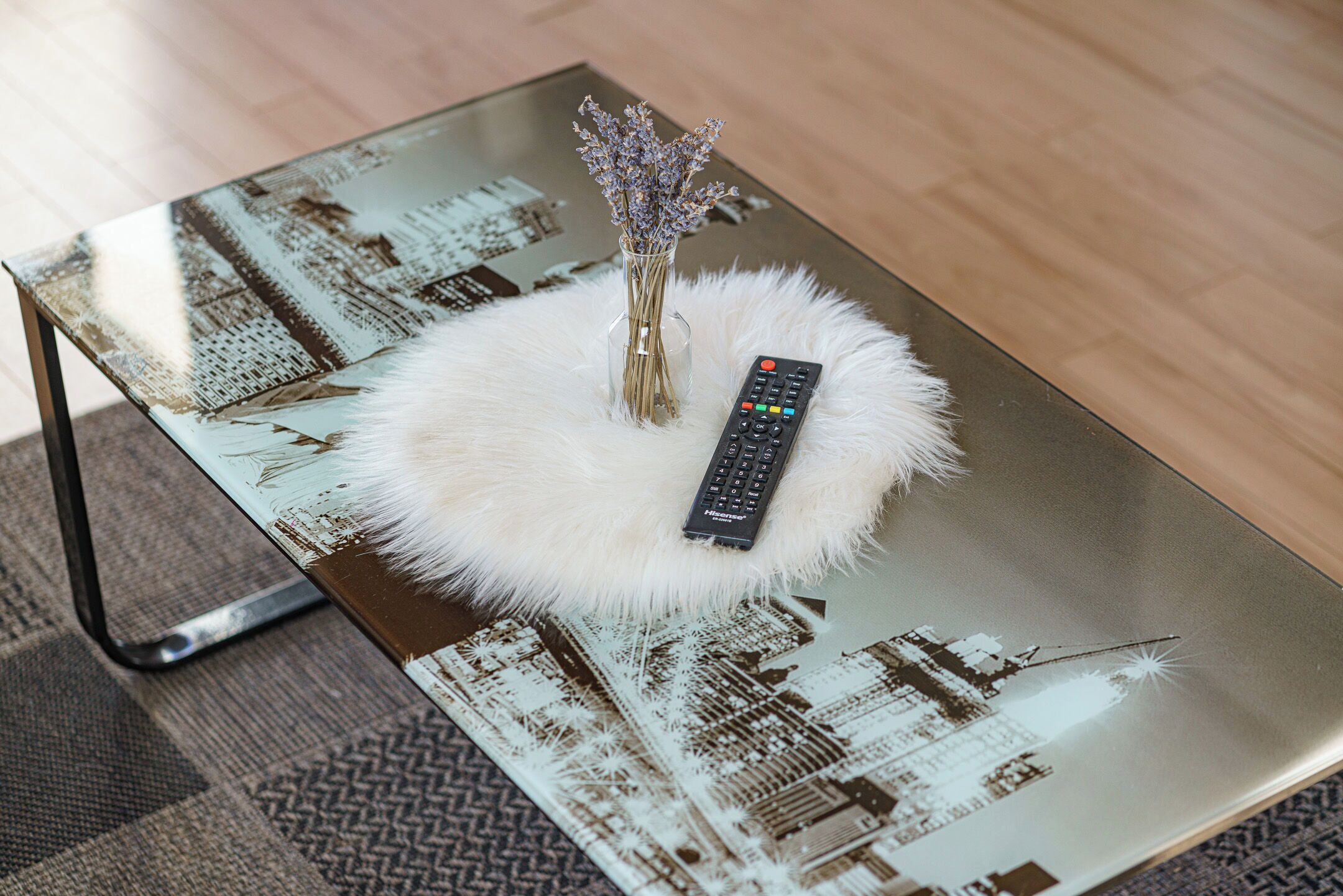 A glass table with a remote control and a vase of flowers on a fluffy white cushion.