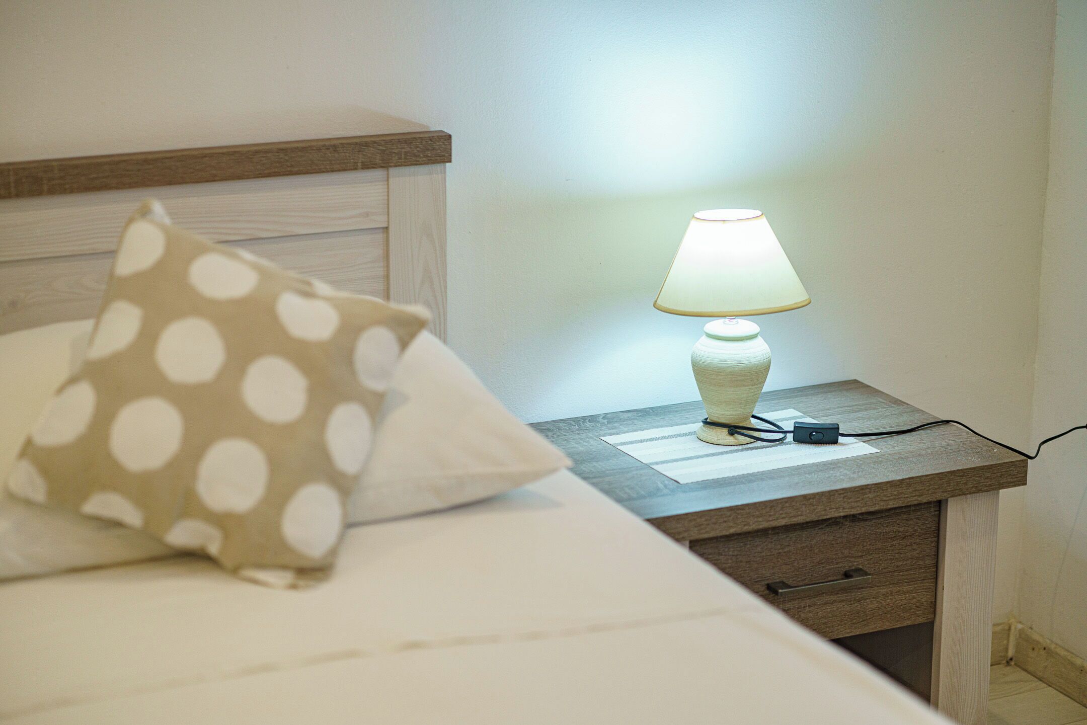 Un lit bien fait, agrémenté d’un coussin à pois, une table de chevet avec une lampe et un tiroir.