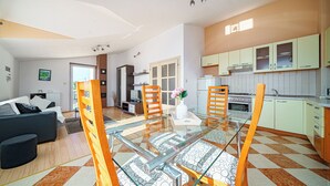 A modern living room with a glass dining table, wooden chairs, and a sofa. There is a kitchen area with light-colored cabinets and a tiled backsplash. - Apartments Zinfandel Vodice (Vodice)