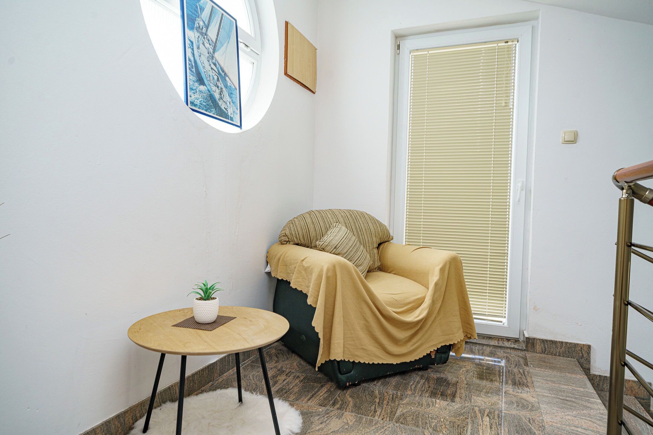 Ein kleiner, gemütlicher Raum mit einem einzelnen grünen Sofa, einem runden Holztisch mit einem Blumentopf und einem großen Fenster mit Jalousien.