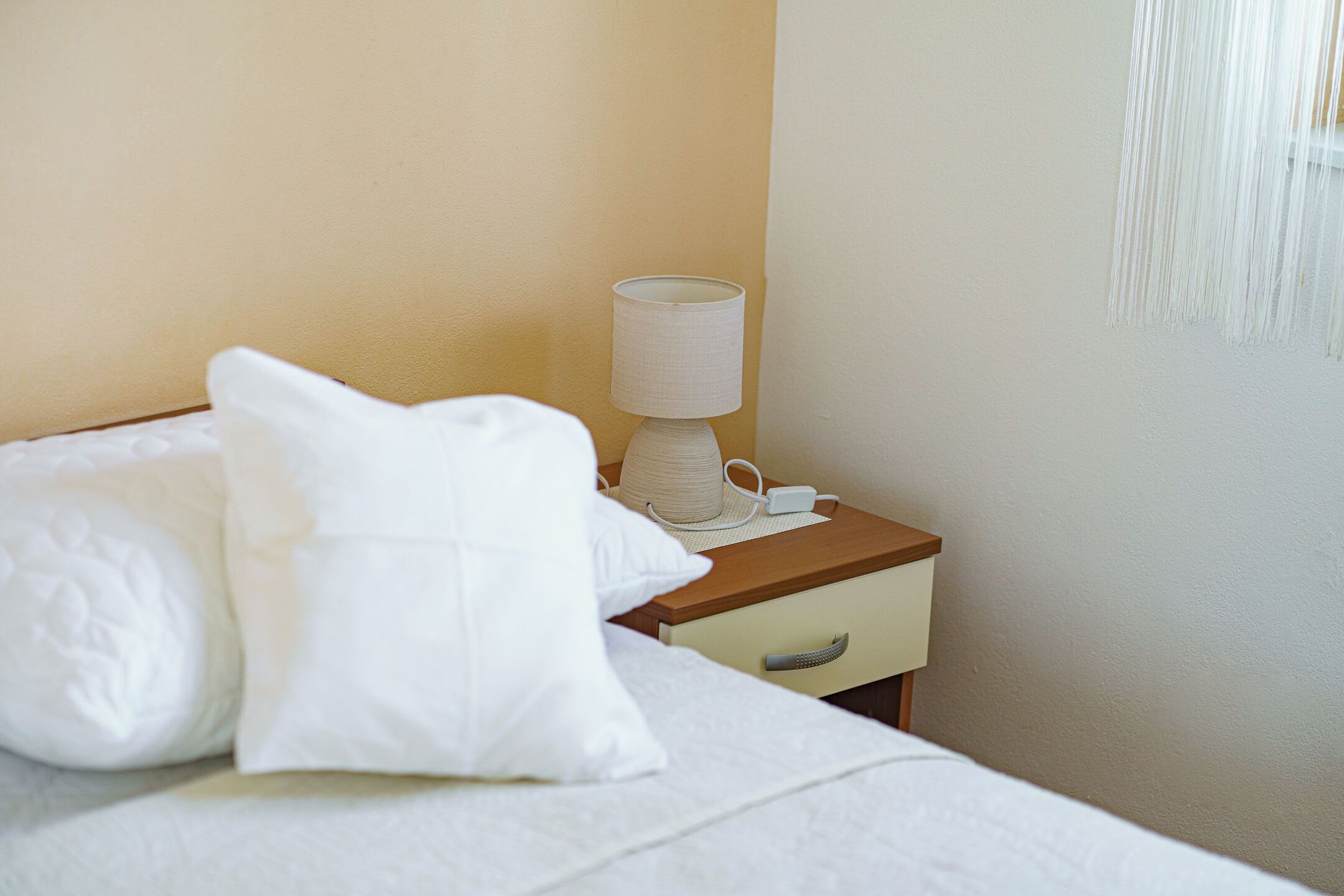Een netjes opgemaakt bed met witte beddengoed en kussens, een nachtkastje met een lamp, en een wand met een macramé wanddecoratie.