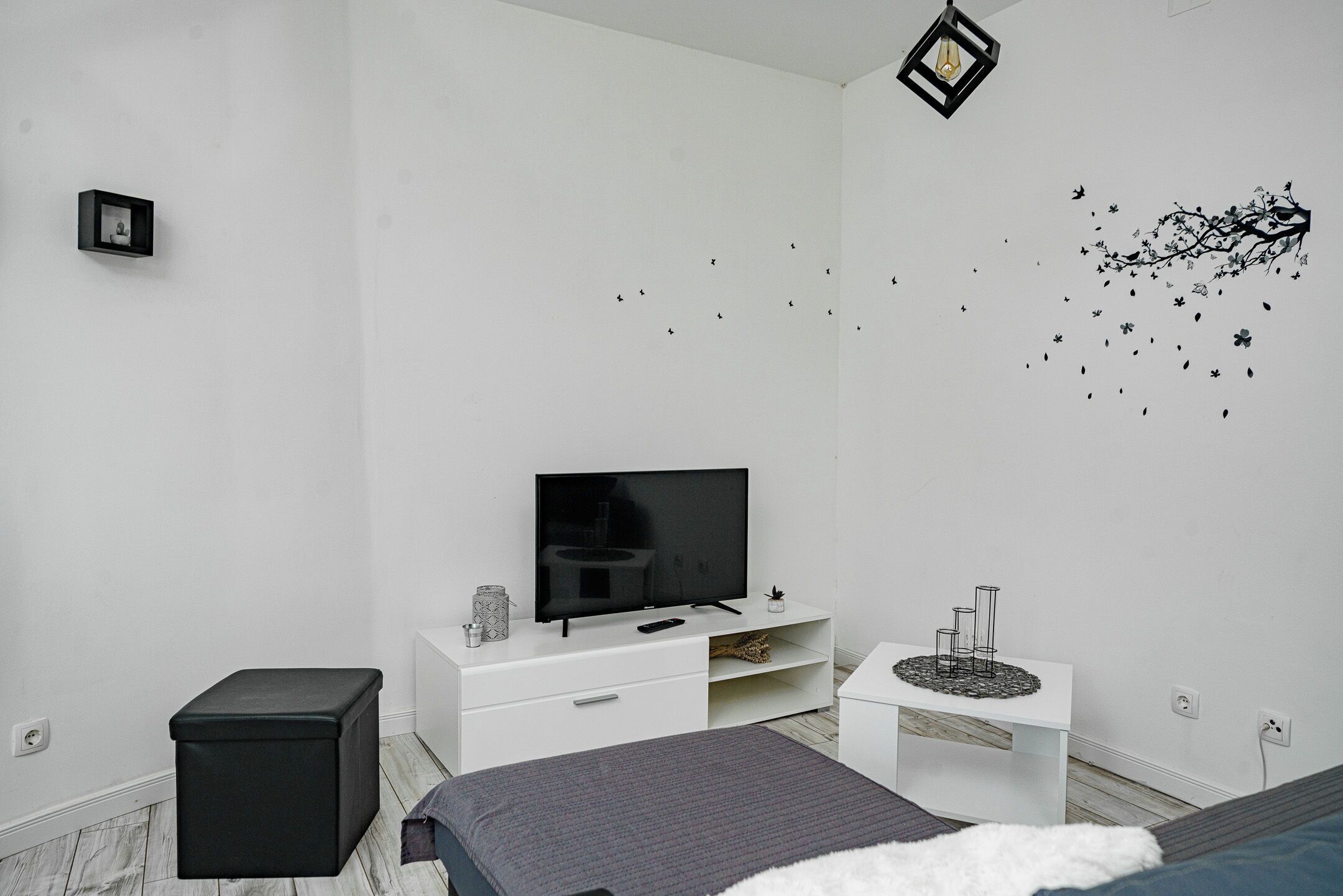Un dormitorio moderno con cama, televisor, cómoda blanca, reposapiés negro y un adhesivo mural.