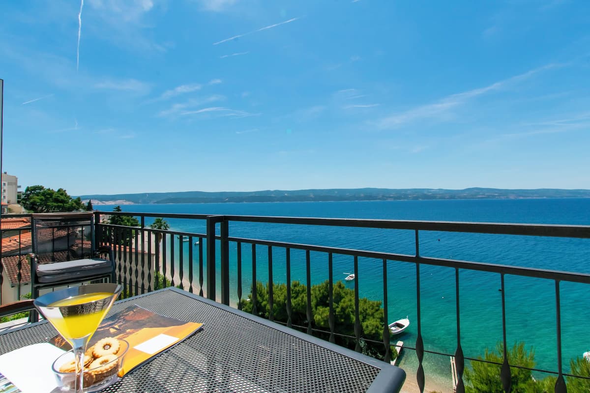 A balcony with a clear view of the sea, a table with a drink and a menu, and a chair.