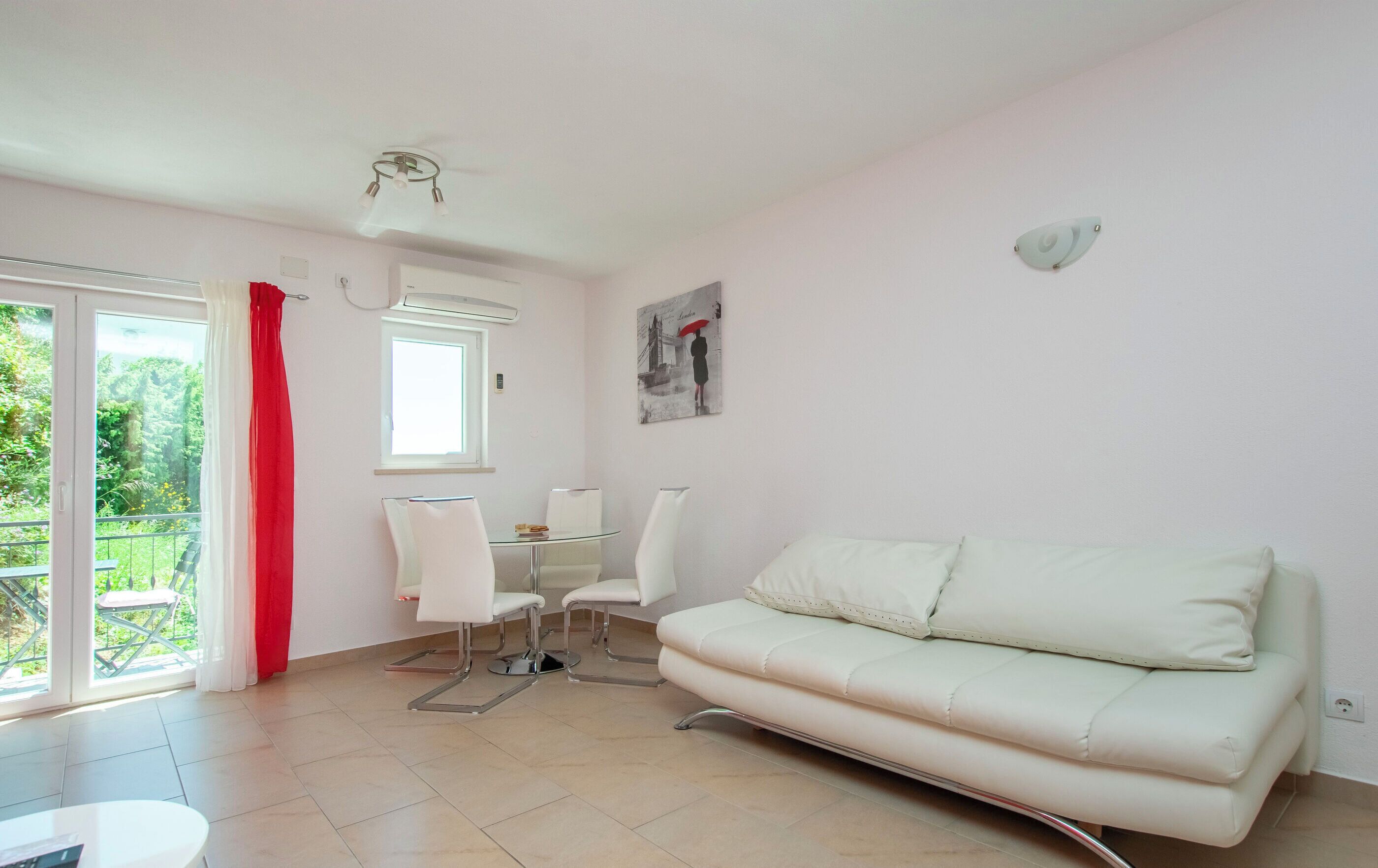 A modern living room with a white sofa, a glass table, and two white chairs. There is a window with a view of greenery outside, a ceiling fan, and a wall-mounted light fixture.