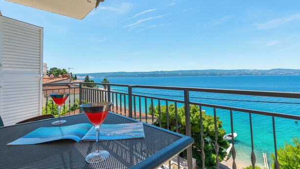 Ein Balkon mit Meerblick, ein Tisch mit einer Zeitschrift und zwei Getränken, sowie ein weißes Gebäude im Hintergrund.