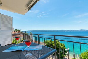 Ein Balkon mit Meerblick, ein Tisch mit einer Zeitschrift und zwei Getränken, sowie ein weißes Gebäude im Hintergrund.