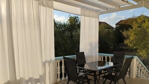 An outdoor patio with a glass table and chairs, white curtains, and a view of greenery and buildings. - Apartment Denis (Sibenik)