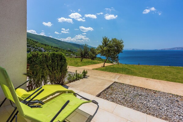 Un balcon avec deux fauteuils de jardin verts, un chemin de gravier et une vue sur la mer et les collines.