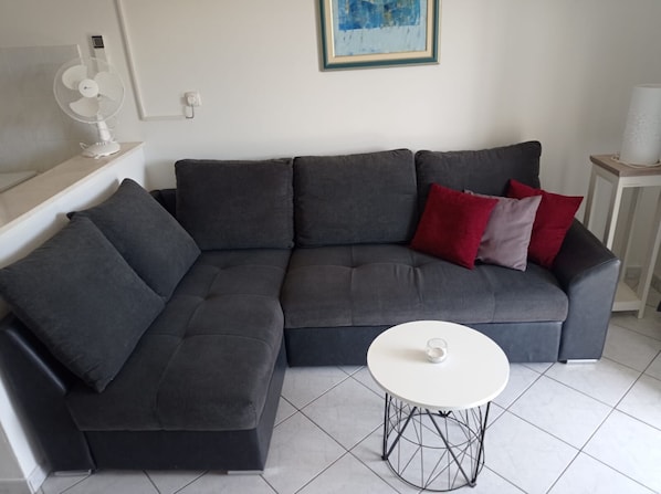 A grey sectional sofa with red and purple cushions, a white table with a glass, and a white fan on a stand. - Apartments Petra (Solta)