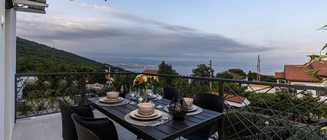 A balcony with a table set for dining, overlooking a scenic view of a coastline and hills.