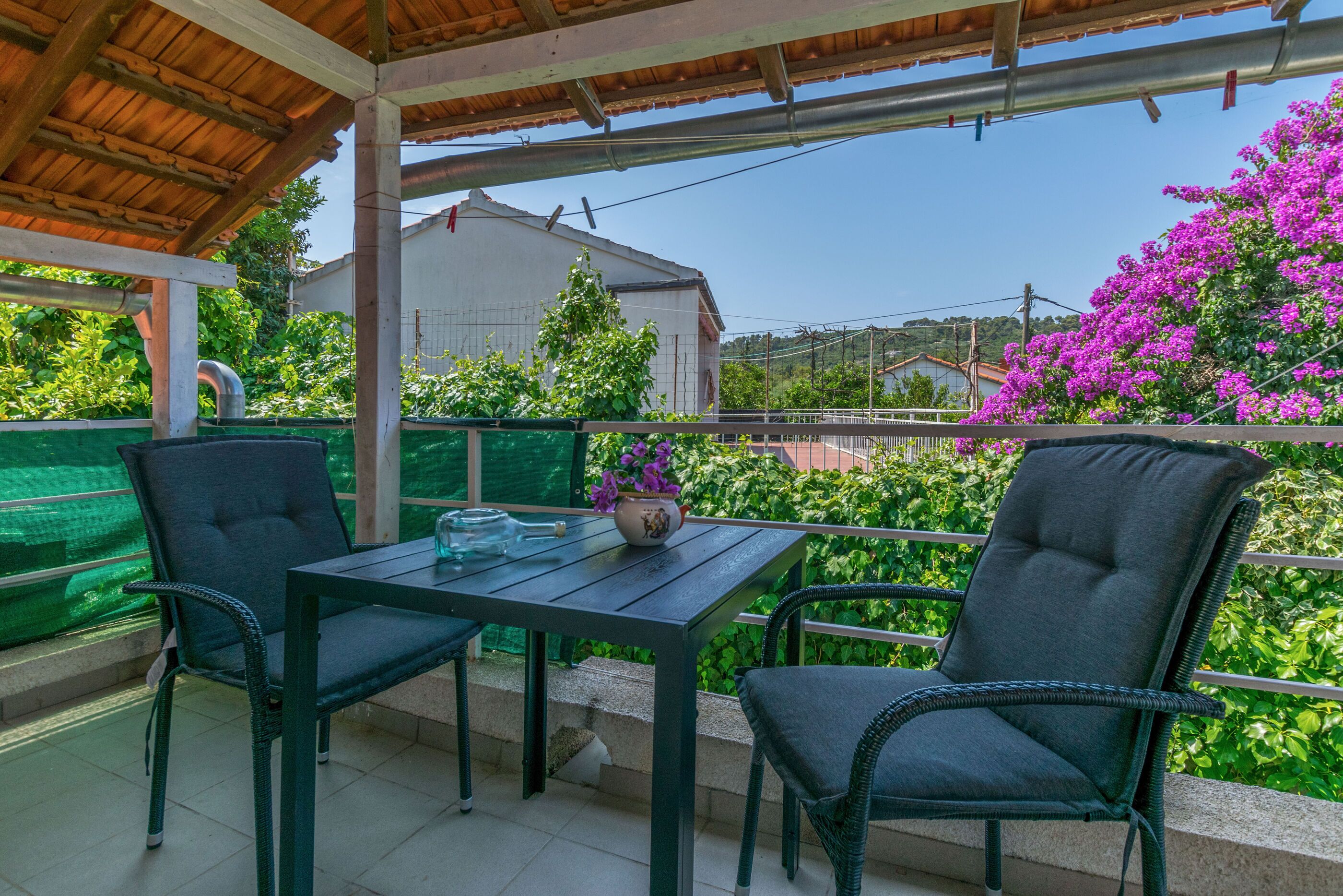Un espace extérieur aménagé, abrité et doté d’une table et de deux chaises, entouré d’une végétation luxuriante et offrant une vue sur un bâtiment blanc et des collines au loin.