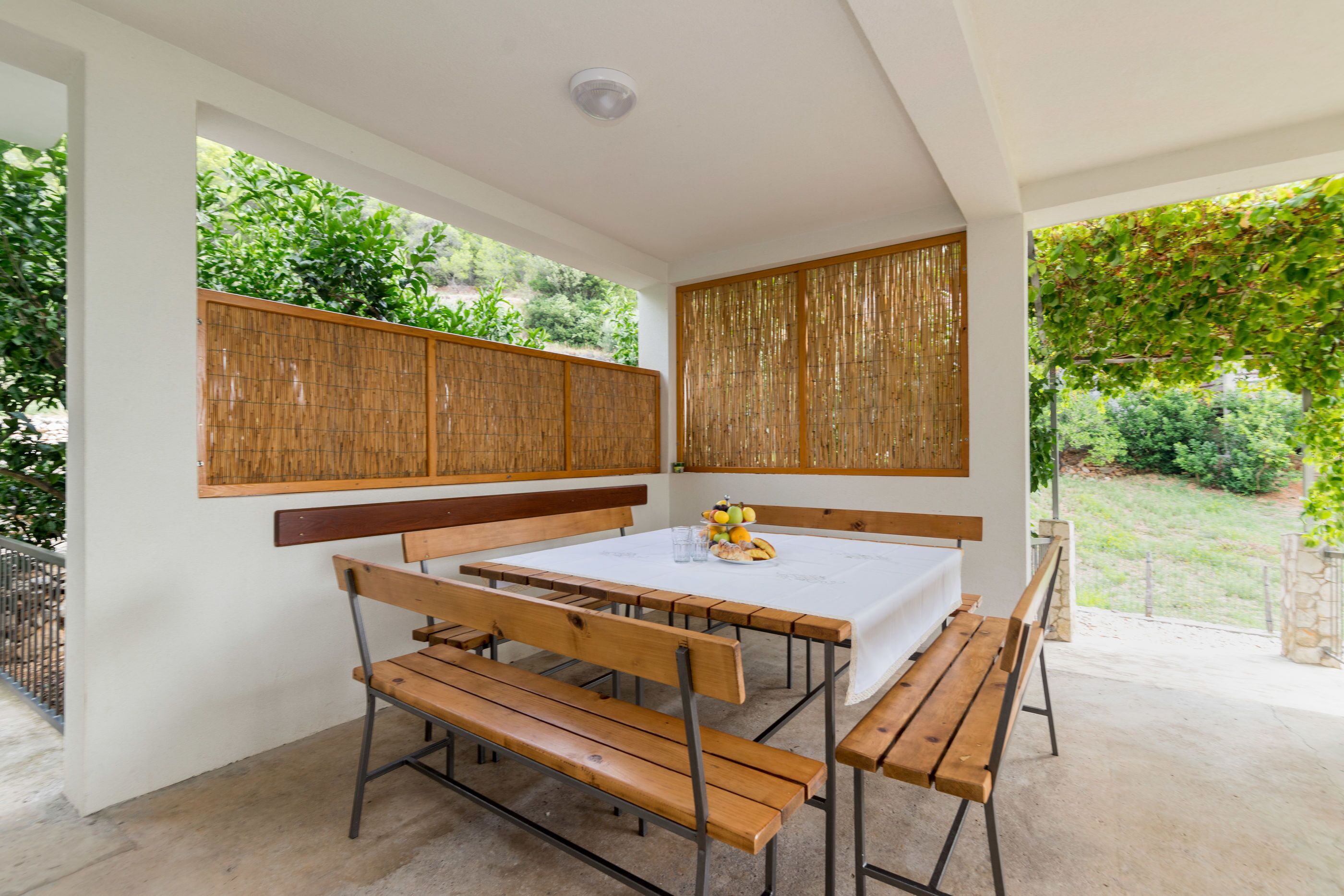 A patio with a wooden table and benches, surrounded by bamboo screens and a view of greenery.