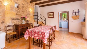 Uma sala de jantar com parede de pedra, mesa de madeira, cadeiras, uma escada e uma cozinha ao fundo.