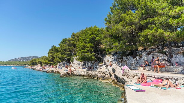 A coastal area with clear blue waters, rocky cliffs, and trees. People are sunbathing and swimming. - House Hacienda (Tisno)