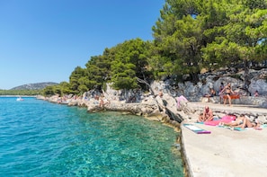 A coastal area with clear blue waters, rocky cliffs, and trees. People are sunbathing and swimming. - House Hacienda (Tisno)