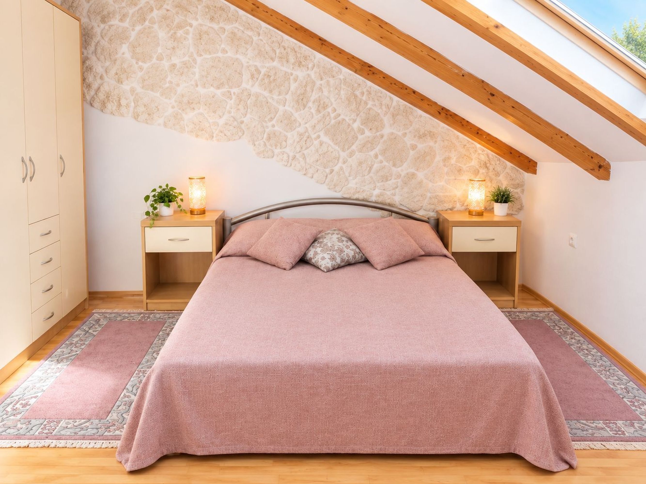A bedroom with a sloped ceiling, a bed with a pink bedspread, two bedside tables with lamps, and a wardrobe.