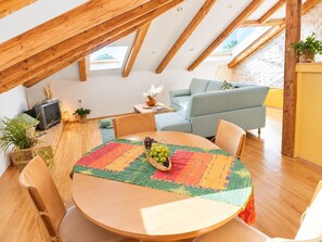 Ein gemütlicher Loftsraum mit Holzbalkendecke, einem runden Esstisch mit farbenfroher Tischläufer und einer bequemen Couch.