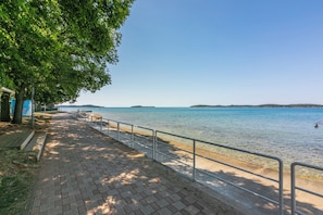 A paved walkway beside a body of water with a metal railing, trees, and distant landforms. - Apartments Radomir (Fazana)