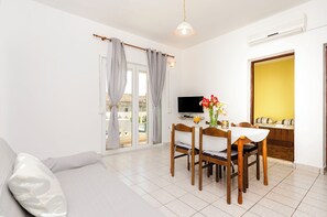Ein modernes Wohnzimmer mit einer Couch, einem Esstisch und Stühlen. Ein Fernseher ist an der Wand befestigt und ein Fenster mit Vorhängen vorhanden.