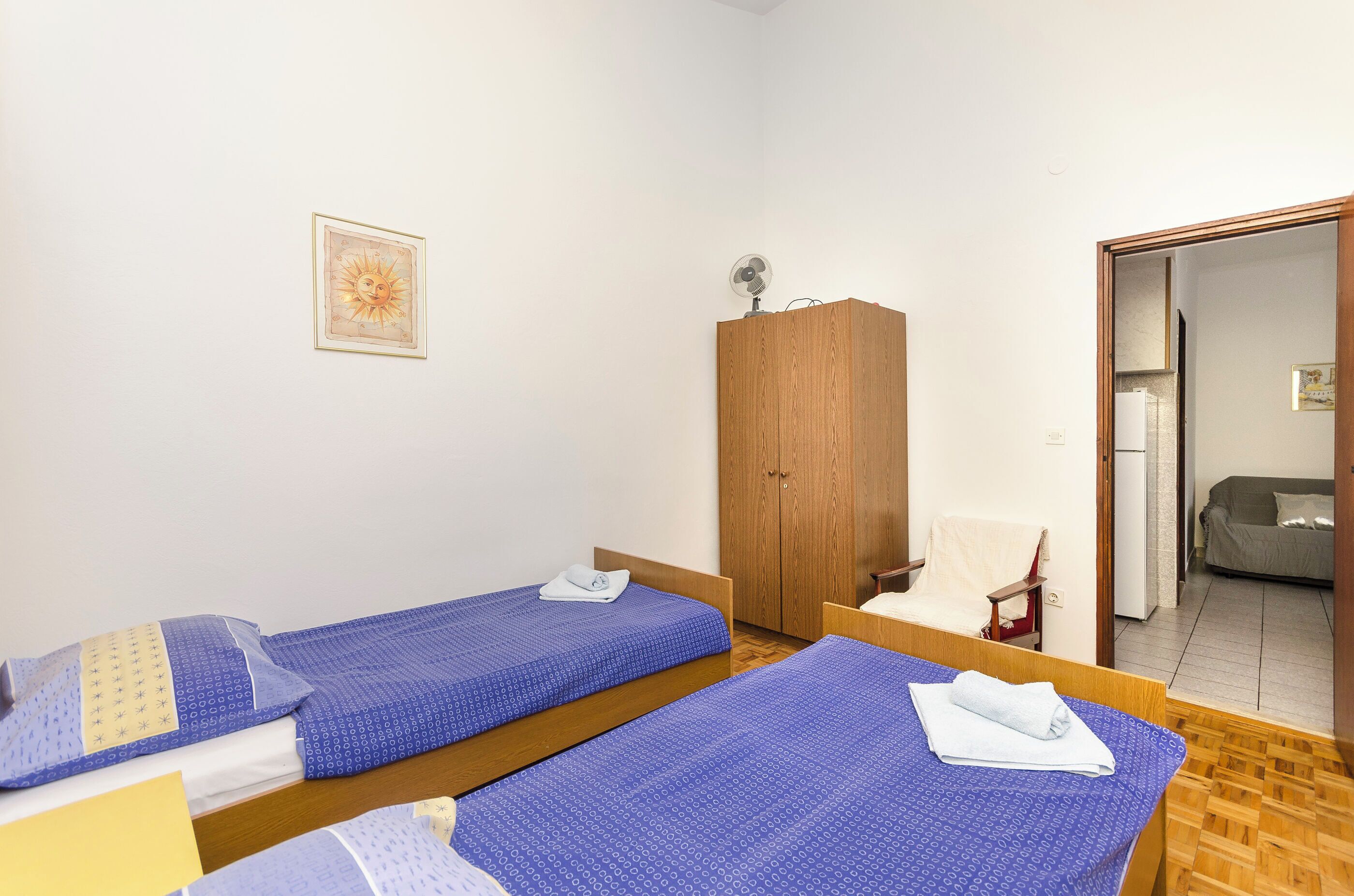 A room with two single beds, a wooden wardrobe, a chair, and a door leading to a living area.