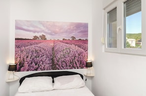 A bedroom with a large bed, a window with blinds, and a wall-mounted picture of a field of flowers. - Apartments and Room Lotus (Tribunj)