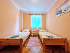 A small, cozy room with two wooden beds, a window with sheer curtains, a small table with a lamp, and a painting on the wall. - Apartment Roses (Novi Vinodolski)
