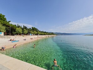 Een strand met helder water, mensen die zwemmen en zonnebaden, en een rij bomen langs de kust.