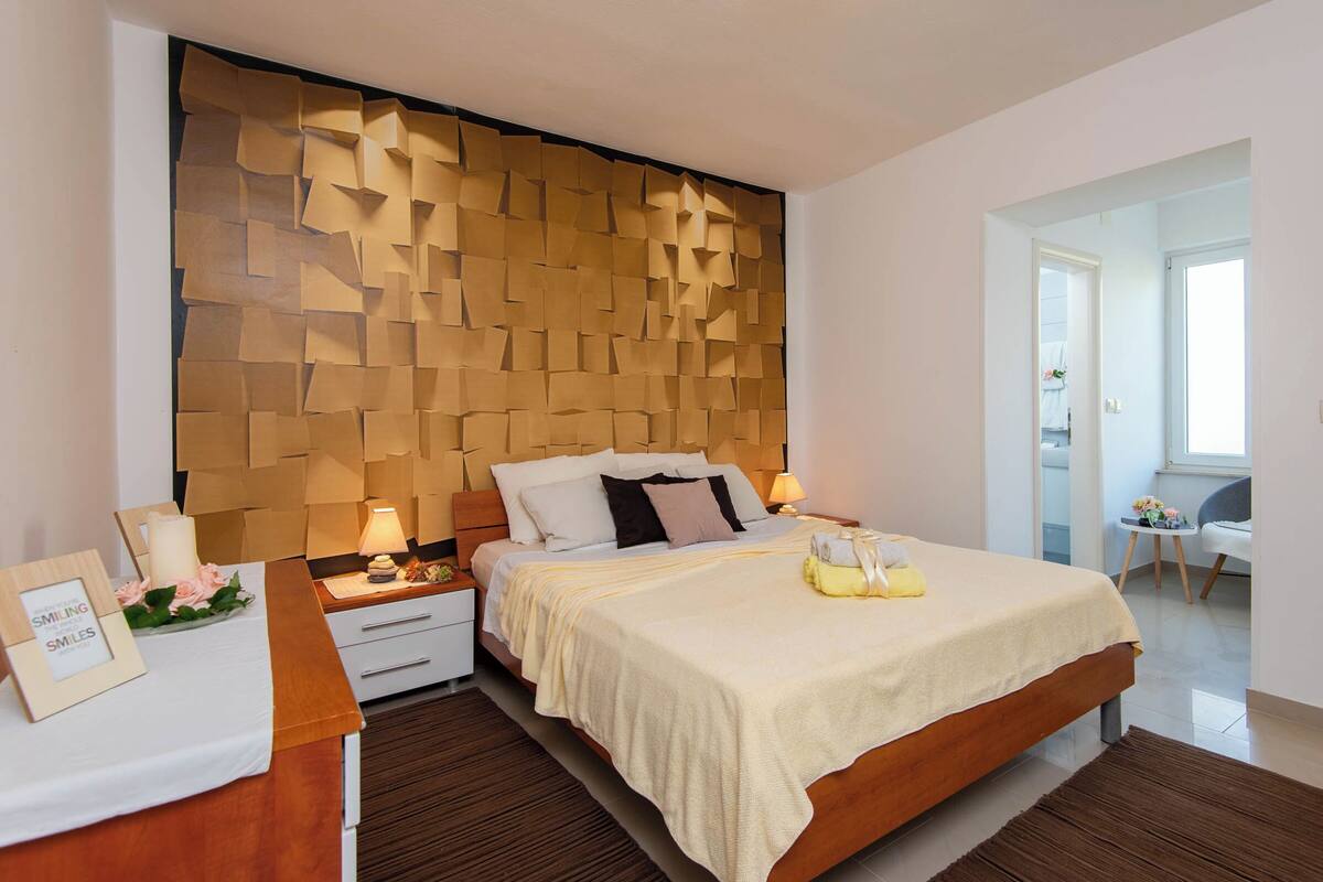 A modern bedroom with a wooden headboard, a large bed with a beige bedspread, bedside tables with lamps, and a small table with a framed picture and flowers.